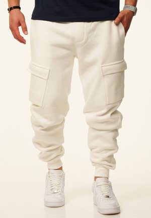 Man wearing white cargo jogger pants with large side pockets and white sneakers, standing with hands partially in pockets.
