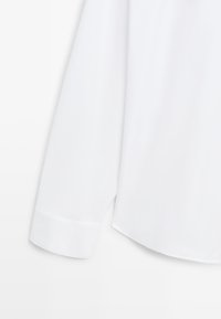 Détail de la manche et du poignet large d'une chemise blanche à manches longues sur un fond blanc uni.