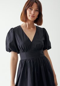 Schwarzes Kleid mit kurzen Puffärmeln, tiefem V-Ausschnitt, tailliertem Schnitt und gerafftem Rock. Hergestellt aus einem leichten Stoff mit glatter Textur.