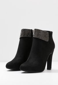 Botas negras de tobillo de ante con un puño de cuentas, presentan una punta afilada y un alto tacón de stiletto. Cierre de cremallera en el lateral.