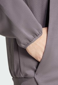 Sivi, lagani materijal s glatkom teksturom; sakupljena manžeta i bočna džep uklopljeni u dizajn odjeće.