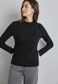 Schwarzes, geripptes Langarmoberteil mit hohem Kragen, im lässigen Schnitt. Kombiniert mit grauen, tapered Hosen mit Gesäßtaschen.