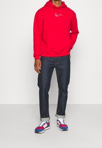Roter Kapuzenpullover mit einer Fronttasche und weißem Logo, kombiniert mit dunkelblauen Jeans und mehrfarbigen Sportschuhen.