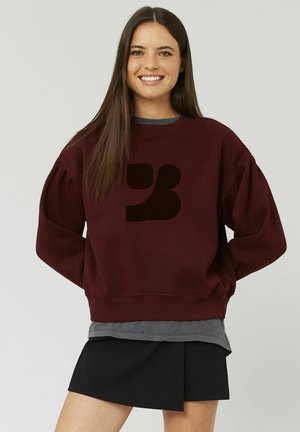Sweatshirt bordeaux avec des manches bouffantes et un logo noir, superposé sur une chemise grise, associé à une jupe plissée noire.