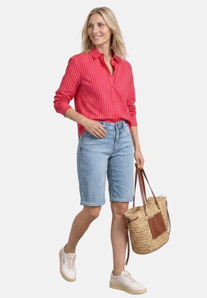 Kvinde iført rødstribet langærmet skjorte og lyse denimshorts, der holder en vævet halmtaske med læderdetaljer.