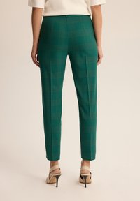 Zielone spodnie w kratę o dopasowanym kroju, smukłym kształcie i gładkiej tkaninie. Posiadają płaski pas i są zestawione z wysokimi obcasami.