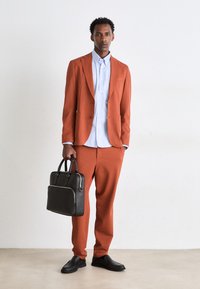 Costume couleur rouille en tissu texturé, associé à une chemise à rayures bleues et blanches à boutons. Sac en cuir noir et chaussures à enfiler.