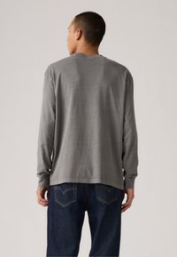 Dlhý rukáv sivá košeľa vyrobená z jemného materiálu, s voľným strihom a okrúhlym výstrihom. Nosená s tmavomodrými džínsami.