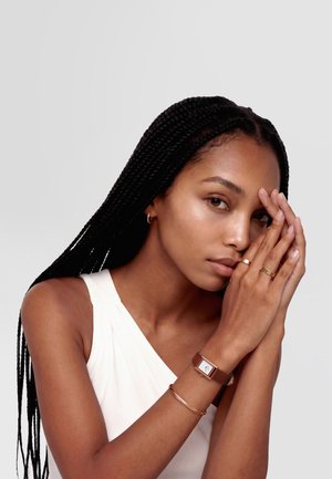 Mlada žena s dugom pletenicom u kosi, nosi bijelu majicu bez rukava, zlatni nakit i sat s smeđim kožnim remenom, ruke blizu lica.