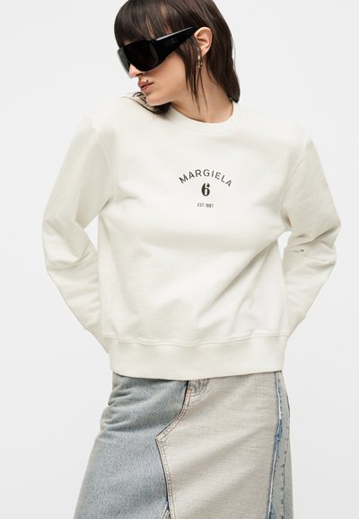 Γυναίκα που φορά υπερμεγέθη μαύρα γυαλιά ηλίου, λευκό φούτερ Margiela και δίχρωμη τζιν φούστα, ποζάροντας με τα χέρια πίσω από την πλάτη.