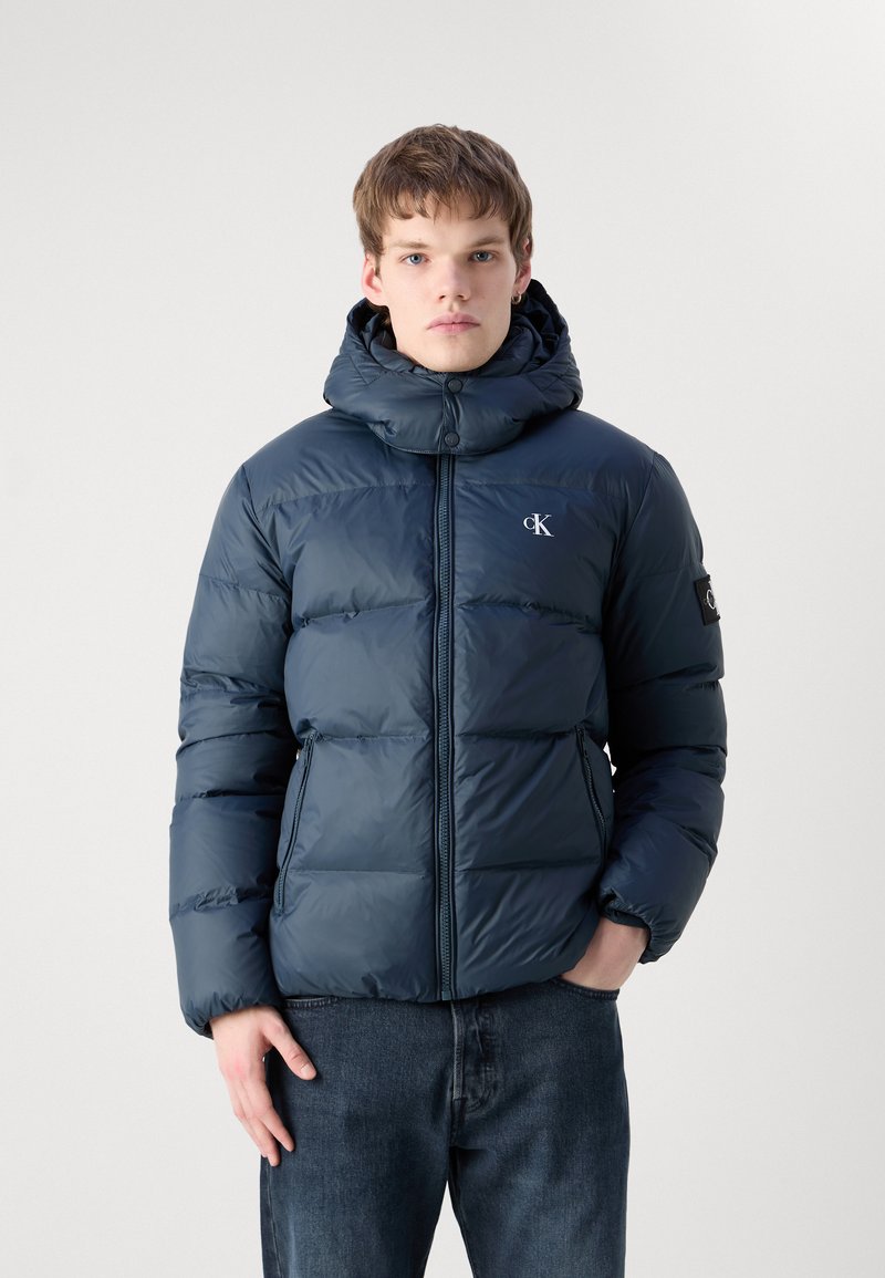 Dunkelblaue Badejacke mit hohem Kragen und Kapuze, mit durchgehendem Reißverschluss, zwei Seitentaschen und Logodetail auf der Brust und dem Ärmel.