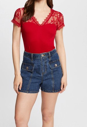 Kobieta ubrana w czerwoną koronkową bluzkę z krótkimi rękawami i dekoltem w serek oraz wysokogatunkowe niebieskie dżinsowe szorty z przednimi kieszeniami z patką.