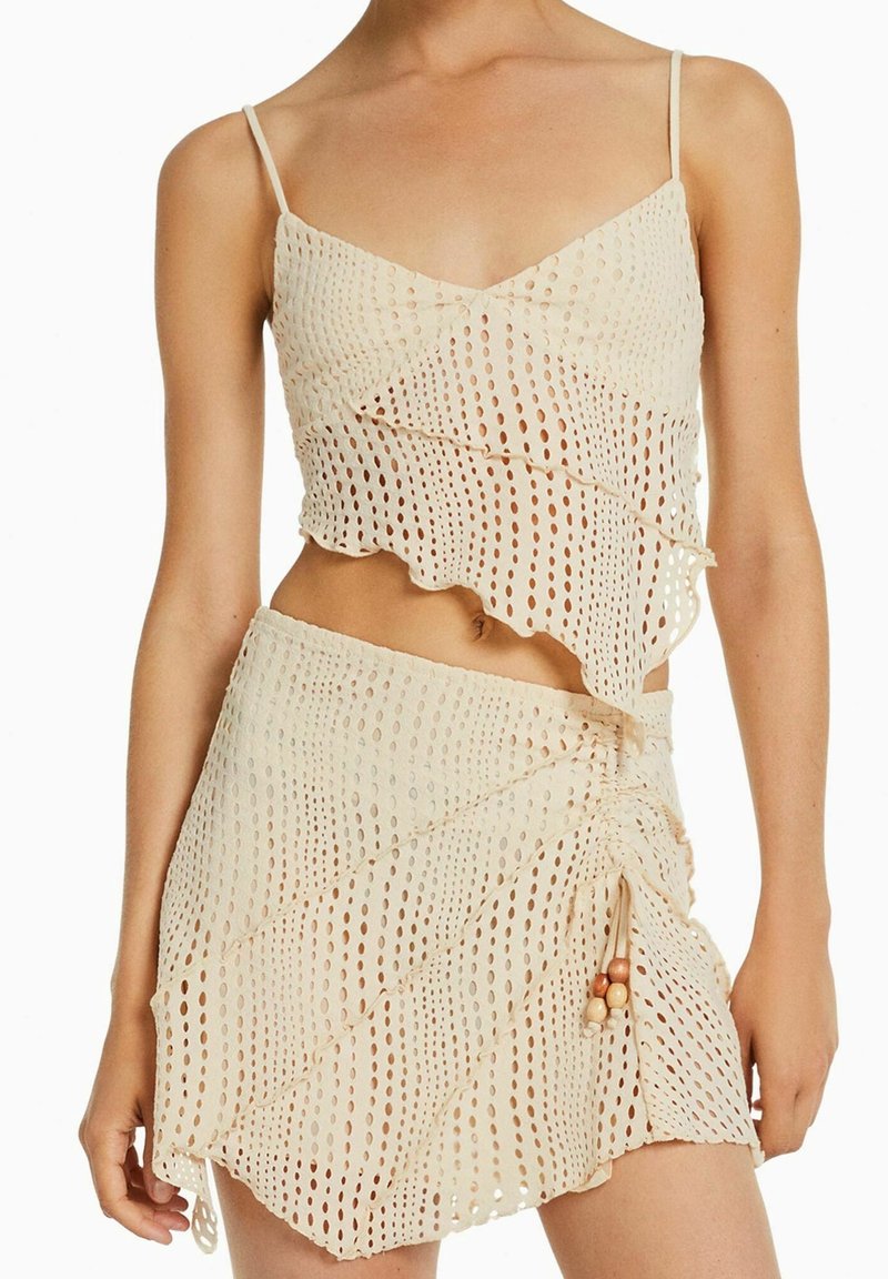 Béžová síťovaná crop top a mini sukně s kulatými výřezy a asymetrickými lemy, s nastavitelnou šňůrkou s dřevěnými korálky na sukni.