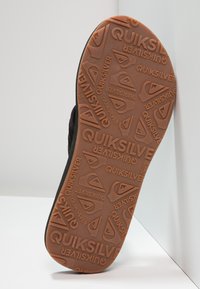 Svart såle med en brun gummibunn som har et mønstrete tekstur og Quiksilver-logoen i ulike posisjoner.