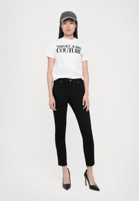 Jauna moteris vilkinti baltą Versace Jeans Couture marškinėlį, juodas kūno prigludusias kelnes, juodus aukštakulnius batus ir pilką beisbolo kepuraitę, stovinti prieš baltą foną.
