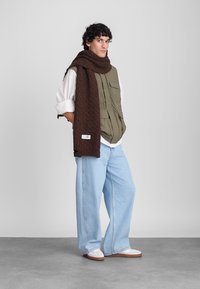 Colete acolchoado verde, camisa branca, lenço castanho estampado, jeans de perna larga azul claro e sapatos brancos com solas castanhas. Design simples e funcional.