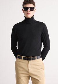 Pulover negru cu guler înalt, confecționat dintr-un material tricotat moale, asortat cu pantaloni khaki și o curea neagră. Modelul poartă ochelari de soare cu rame mari.