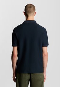 Marineblå polo-shirt med struktureret stof, korte ærmer og flad krave, parret med grønne bukser. Bagfra ses designdetaljerne.