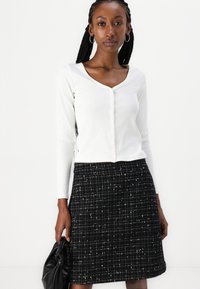 Top blanc à manches longues côtelé avec un col en V et un détail de bouton, associé à une jupe en tweed noir arborant un motif à carreaux et des accents mouchetés.