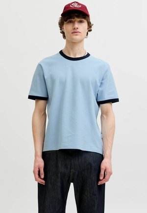 Jeune homme portant un t-shirt bleu clair avec une bordure noire, un jean foncé et une casquette de baseball rouge sur un fond blanc uni.