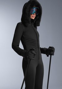 Czarny kombinezon narciarski z kapturem, ozdobiony sztucznym futrem, o ribbowanej strukturze, z rękawiczkami z dziurkami na kciuki, trzymający w obu rękach kije narciarskie.