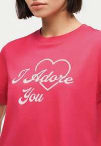 Γυναίκα που φοράει ένα φουξια μπλουζάκι με λευκό κείμενο «Σε λατρεύω» και περίγραμμα καρδιάς στο στήθος, με κομμένο κάτω από τη μύτη.