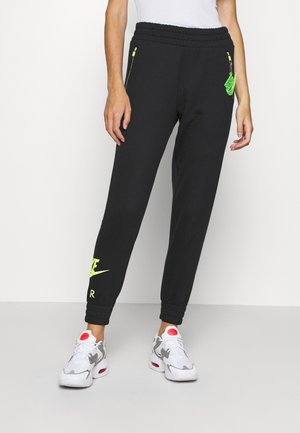 Person trägt schwarze Nike-Sweatpants mit neongrünem Logo und weiße Sneakers mit pinken Akzenten, steht vor einem schlichten weißen Hintergrund.