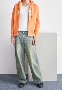 Felpa arancione con zip e logo bianco, indossata sopra una canotta bianca; abbinata a jeans a gamba larga, sbiaditi di colore blu, e scarpe slip-on nere.
