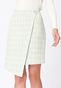 Jupe portefeuille tissée vert clair avec un ourlet asymétrique et une fermeture à un seul bouton, portée avec un haut blanc à manches longues.