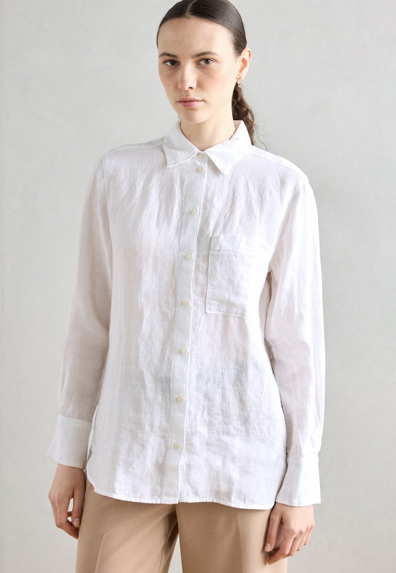 Camicia button-up bianca in lino con maniche lunghe, colletto, taschino sul lato sinistro e orlo leggermente curvato. Texture liscia.