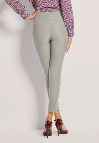 Frau in hellgrauen, figurbetonten Hosen, bordeauxfarbenem, gemustertem Langarmblusenshirt und bordeauxfarbenen High Heels, steht mit einer Hand auf der Hüfte.