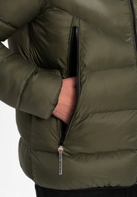 Olivgrön puffjacka med ett kviltat mönster, som har en svart dragkedjeficka och en silverdragare för enkel åtkomst. Mjuk textur.