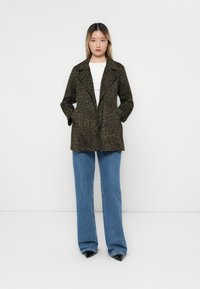 Olivgrön dubbelknäppt jacka med leopardmönster, med stora slag och sidofickor, matchad med ljusa denimbyxor med utsvängda ben.