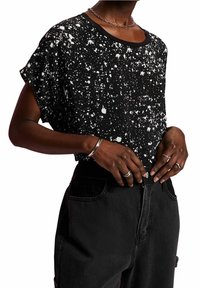 Camisa negra de manga corta con patrón de salpicaduras blancas, escote redondo, combinada con unos jeans oscuros de cintura alta. Se pueden ver anillos y una pulsera de plata como acentos.