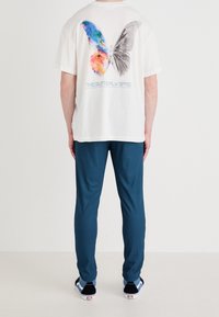 Kremowa koszulka z kolorowym graficznym nadrukiem motyla na plecach, zestawiona z ciemnoniebieskimi dresami i czarnymi sneakersami z niebieskimi akcentami.