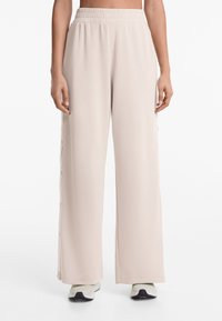 Helle beige weit geschnittene Hose aus weichem Stoff mit elastischem Bund und seitlichen Knöpfen für eine verstellbare Passform. Strukturiert, glatte Oberfläche.