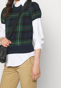 Top de cuadros navy sin mangas con detalles en verde, rojo y amarillo, combinado con una camisa blanca de manga larga. Bolso de hombro con cuero negro texturizado.
