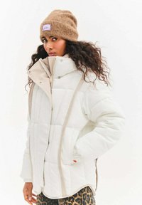 Biała kurtka puchowa z beżowymi akcentami i poziomym pikowaniem, zestawiona z brązową dzianą czapką z łatką z logo w jasnym fiolecie.