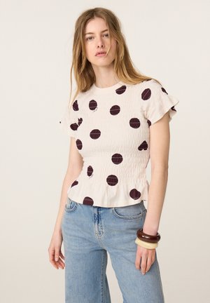 Young woman wearing a white short-sleeve top with large black polka dots, light blue jeans, and chunky bracelets on her right wrist.