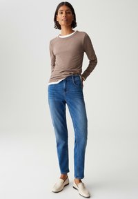 Persoon die staat met een bruin langemouwenshirt, blauwe jeans, witte onderhemd en beige instappers tegen een effen achtergrond.