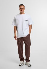 Witte katoenen T-shirt met korte mouwen, met een zwart bedrukte logo op de linkerkant van de borst. Gekocht met een paar bruine broeken en zilveren sportschoenen.