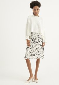 Blusa bianca corta con scollatura a volant e maniche voluminose, abbinata a una gonna a fiori in bianco e nero lungo fino al ginocchio. Tacchi di colore chiaro.