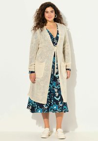 Cardigan lungo a maglia color crema con chiusura a nastro, dotato di due tasche laterali, indossato sopra un vestito floreale blu navy con motivi blu e bianchi.