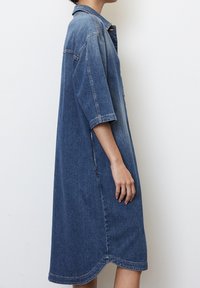 Sukienka koszulowa z denimu, o długości średniej, z kołnierzykiem, krótkimi rękawami i bocznymi rozcięciami. Jasnoniebieski odcień, miękka faktura i widoczne detale przeszyć.