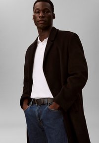 Fekete kabát sima textúrával, fehér galléros póló, kék farmernadrág és fekete öv. A modell zsebre tett kézzel áll.