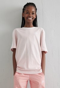 Camiseta de manga corta rosa claro con el logo blanco de adidas en el pecho, combinada con pantalones rosa claro a juego. Textura de tela suave.