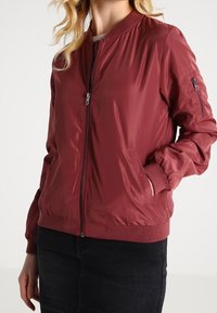 Bordový bomberový kabát, nylonová tkanina, zipová léga, žebrované manžety a lem, postranní kapsy a jedna kapsa na zip na rukávu. Hladká, lesklá textura.