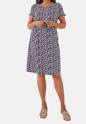 Vrouw draagt een knie-lange marineblauwe jurk met wit en rood bloemmotief, korte mouwen, gestrikte taille en beige instapschoenen.