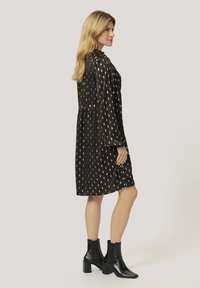 Vestito nero con motivo a foglia dorata, colletto alto, maniche lunghe e vita arricciata. Abbinato a stivaletti neri fino alla caviglia con tacco a blocco.