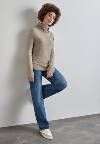 Beige pletený svetr s vysokým límcem, spárovaný s modrými širokými džíny. Bílé tenisky s krémovými akcenty. Pozadí je neutrální.